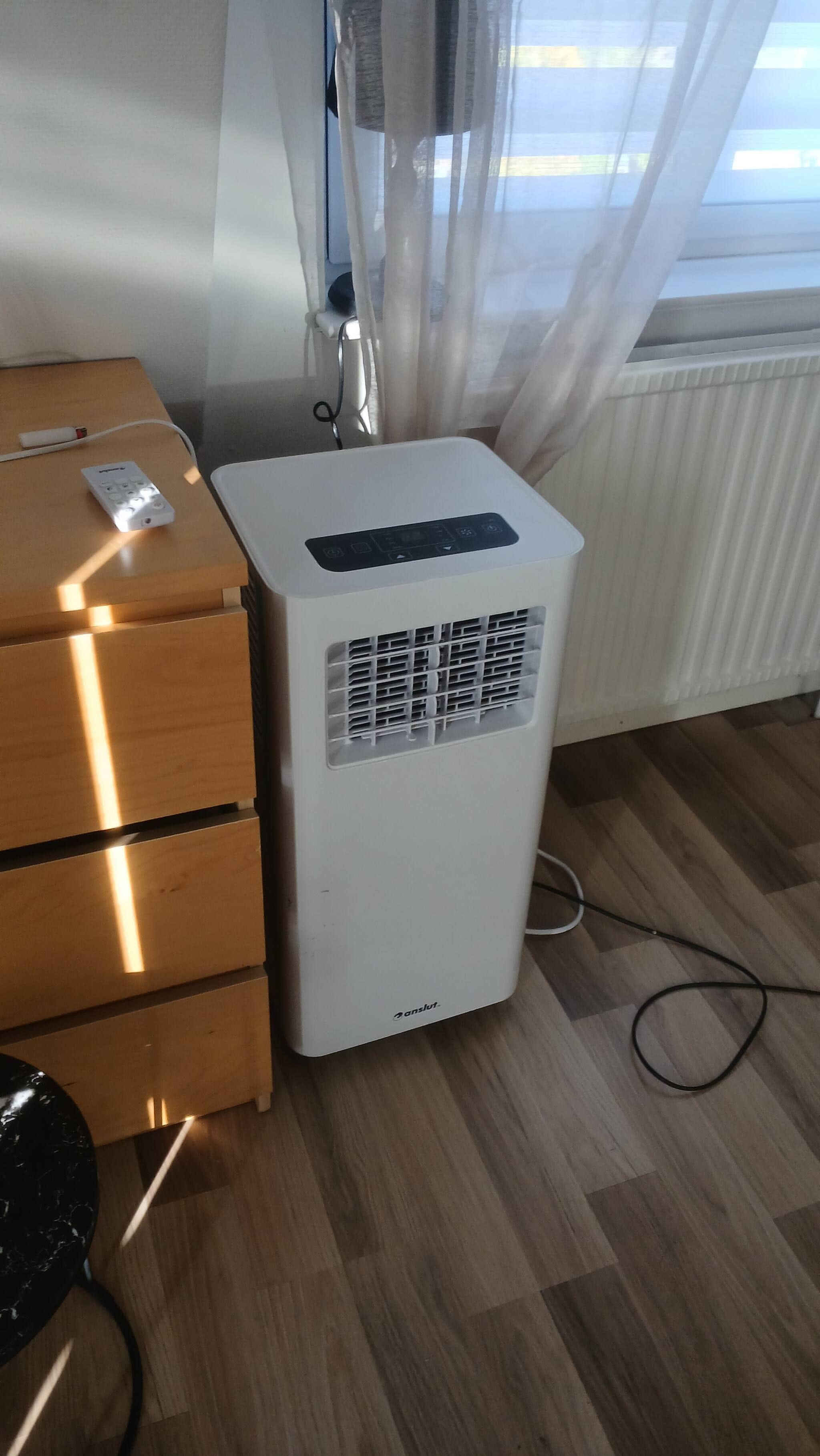En hvid aircondition-enhed med kontrolpanel, en trækommode og en radiator i et rum med trægulv.