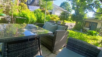 An outdoor patio with wicker furniture, a glass table, and a view of a garden and buildings in the background.
