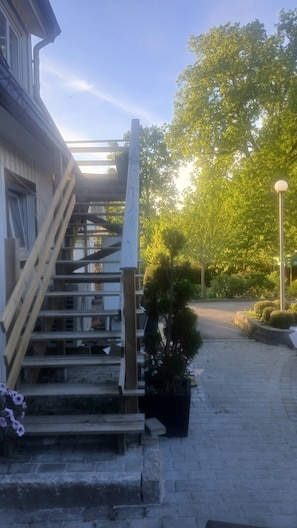 Un edificio con una escalera que lleva a una zona cubierta, rodeado de vegetación y un sendero pavimentado.