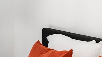 Een bed met een zwart hoofdeinde, witte en oranje kussens, en een klein rond tafeltje met een zwart oppervlak.