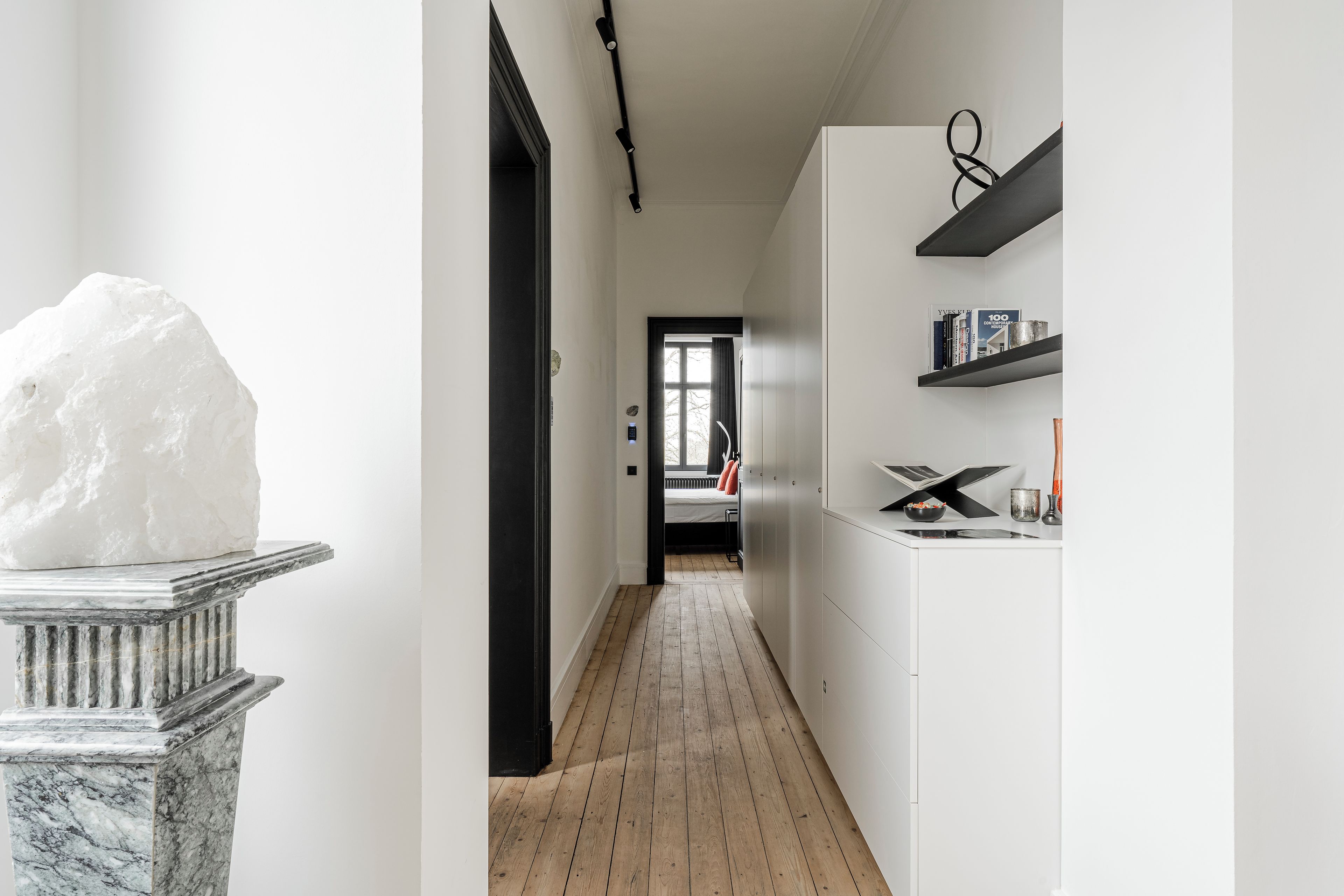 Un couloir doté d’un piédestal en marbre, d’un plancher en bois, de murs blancs et d’une porte donnant sur une autre pièce.