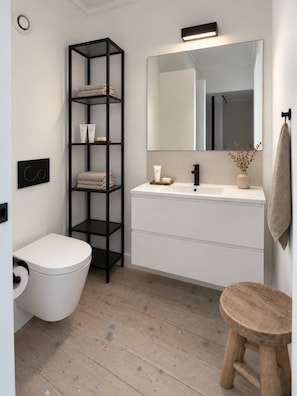 A modern bathroom with a white toilet, a black shelving unit with towels, a white sink with a mirror, and a wooden stool. - Boutique Townhouse Suite | Bruges Centre (Brugge)