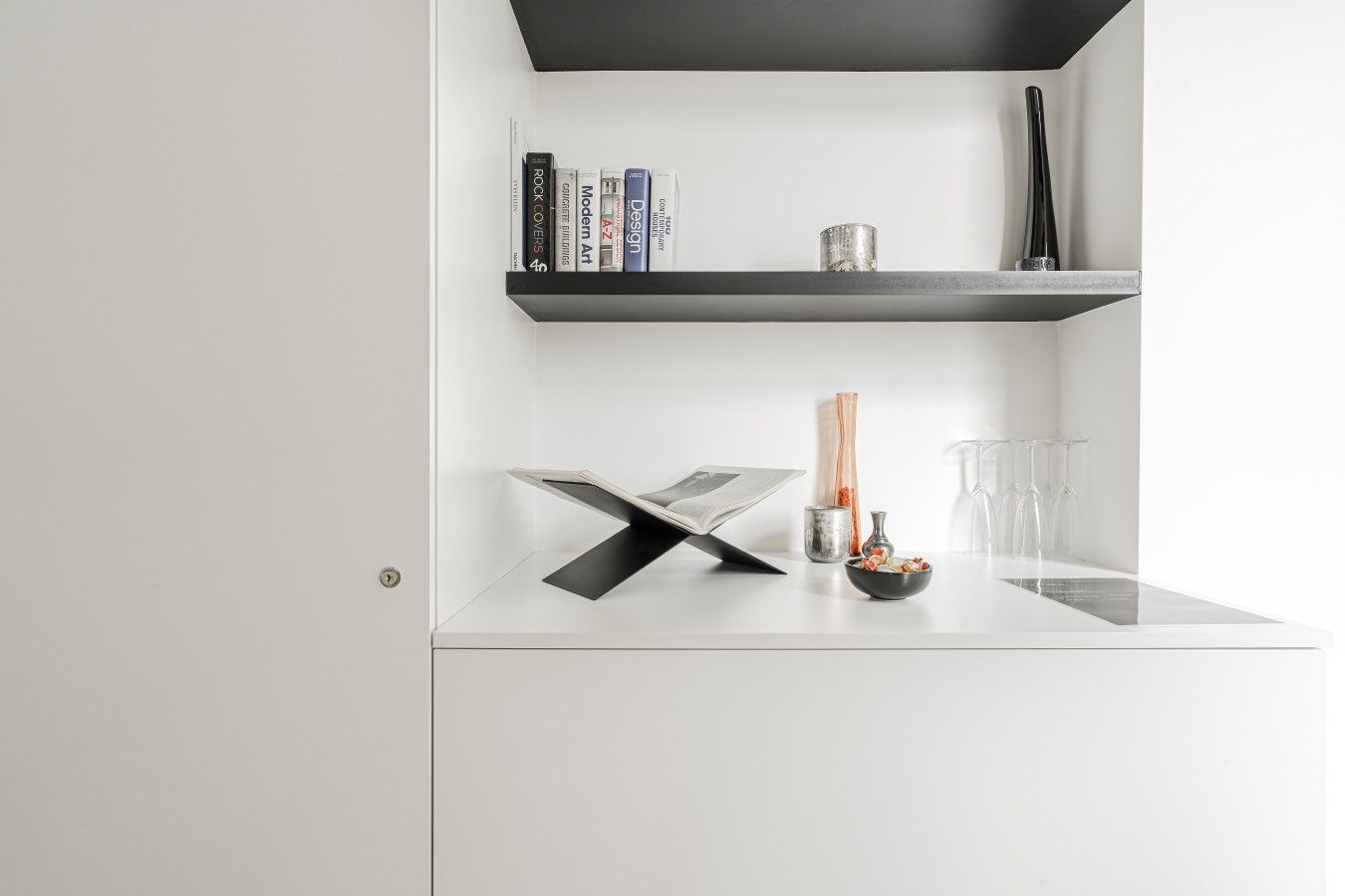 Une pièce moderne et minimaliste, avec un bureau blanc, une étagère noire contenant des livres, un vase et un pichet en verre.