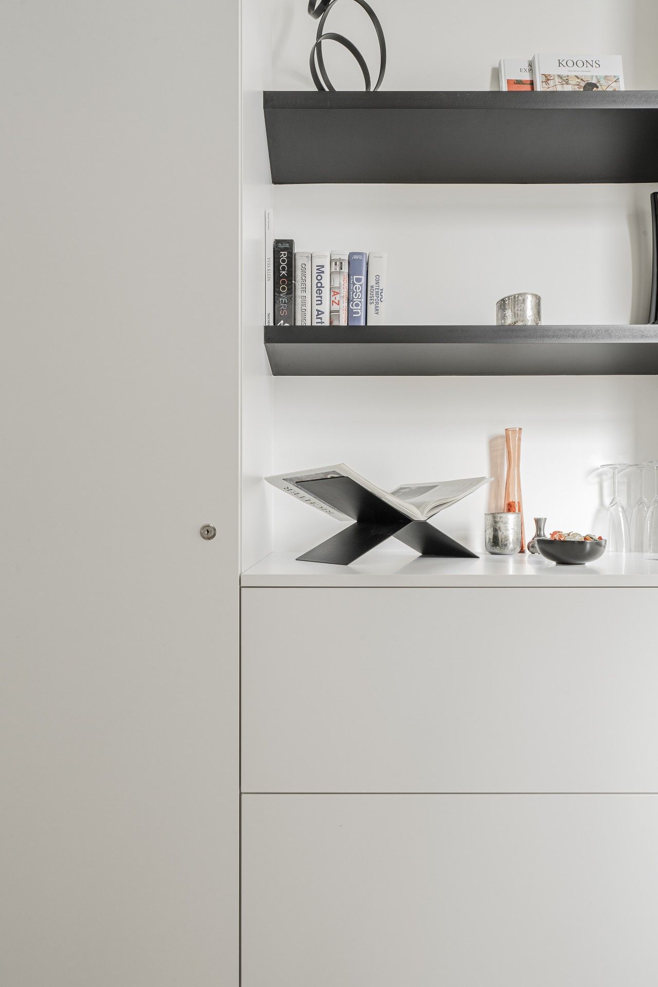 Un interno moderno con una parete bianca, due mensole nere e un tavolo minimalista con un libro, un vaso e una ciotola.