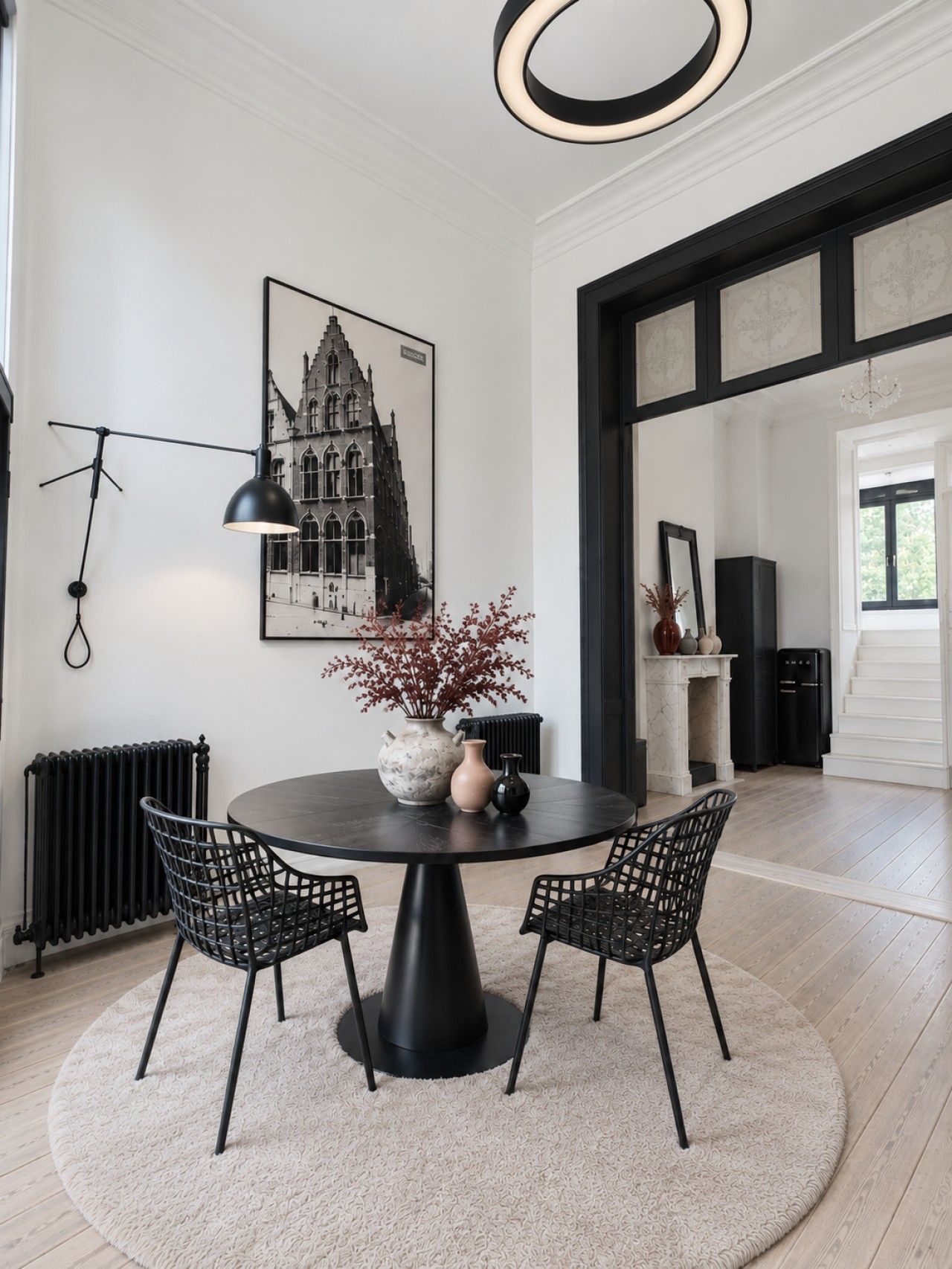 Een eetgedeelte met een ronde tafel, twee stoelen, een vaas met bloemen, een zwart hanglamp en een ingelijst schilderij aan de muur.