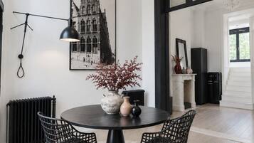 Een eetgedeelte met een ronde tafel, twee stoelen, een vaas met bloemen, een zwart hanglamp en een ingelijst schilderij aan de muur.