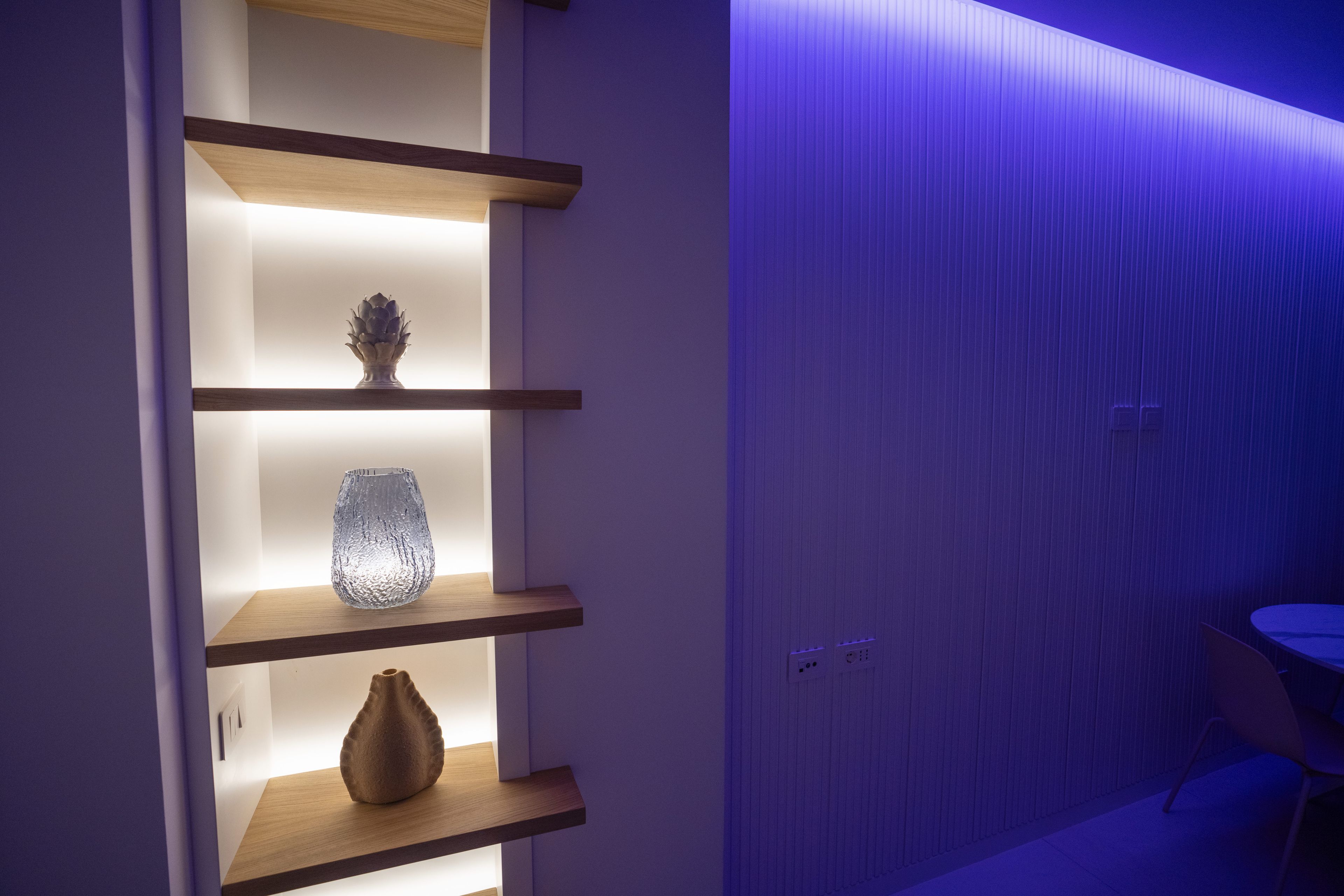 Un intérieur moderne avec des étagères en bois où sont exposés des objets décoratifs, un mur orné de lignes verticales et une table avec des chaises.