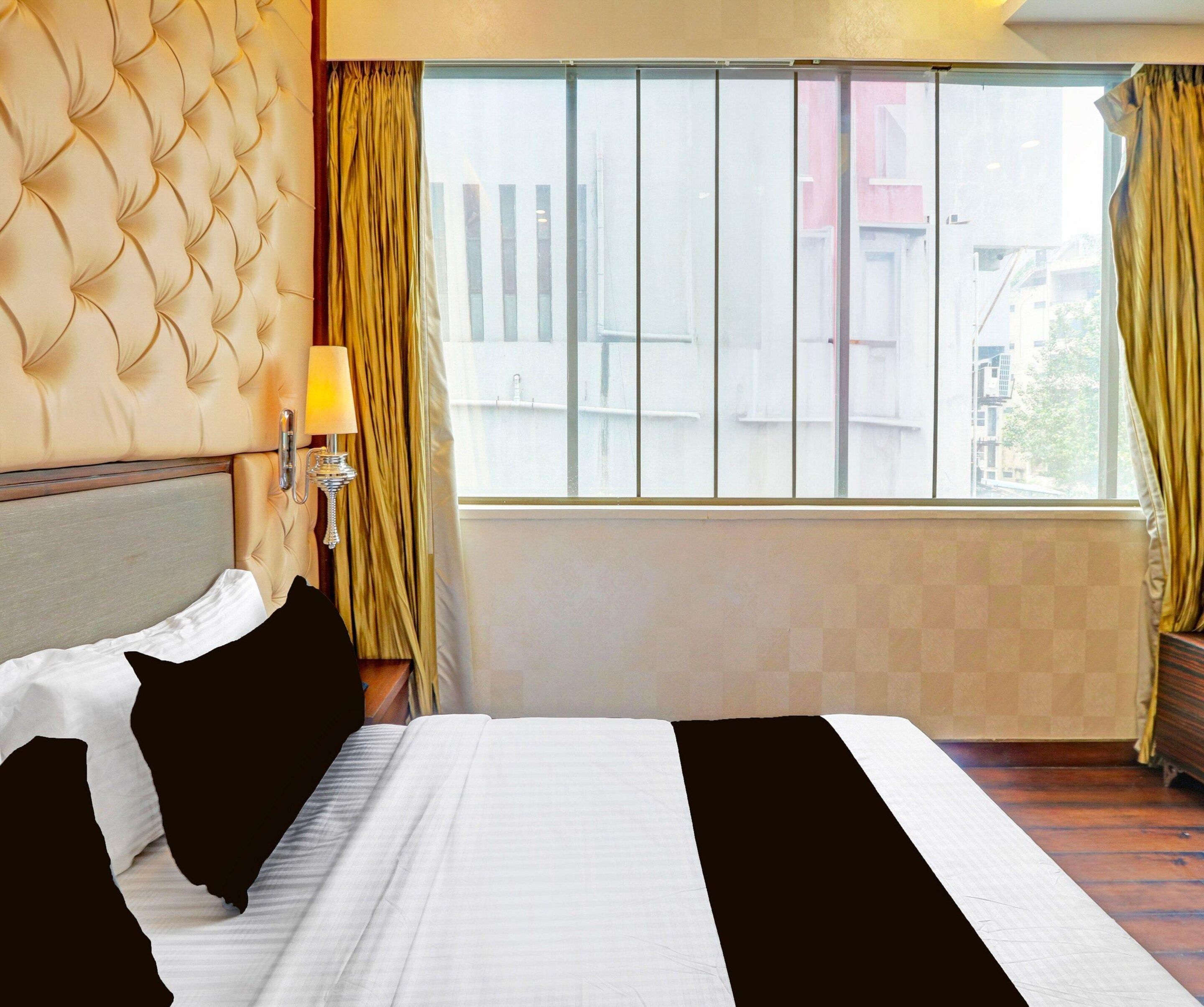 Habitació d'hotel amb un llit, una tauleta de nit, una làmpada i una finestra amb cortines.