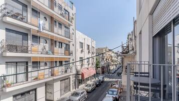 A narrow urban street flanked by multi-story residential buildings with balconies, parked cars, and a clear sky.