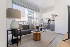 A modern apartment with a sofa, a round wooden side table, a lamp, and a kitchen area with a refrigerator and cabinets.