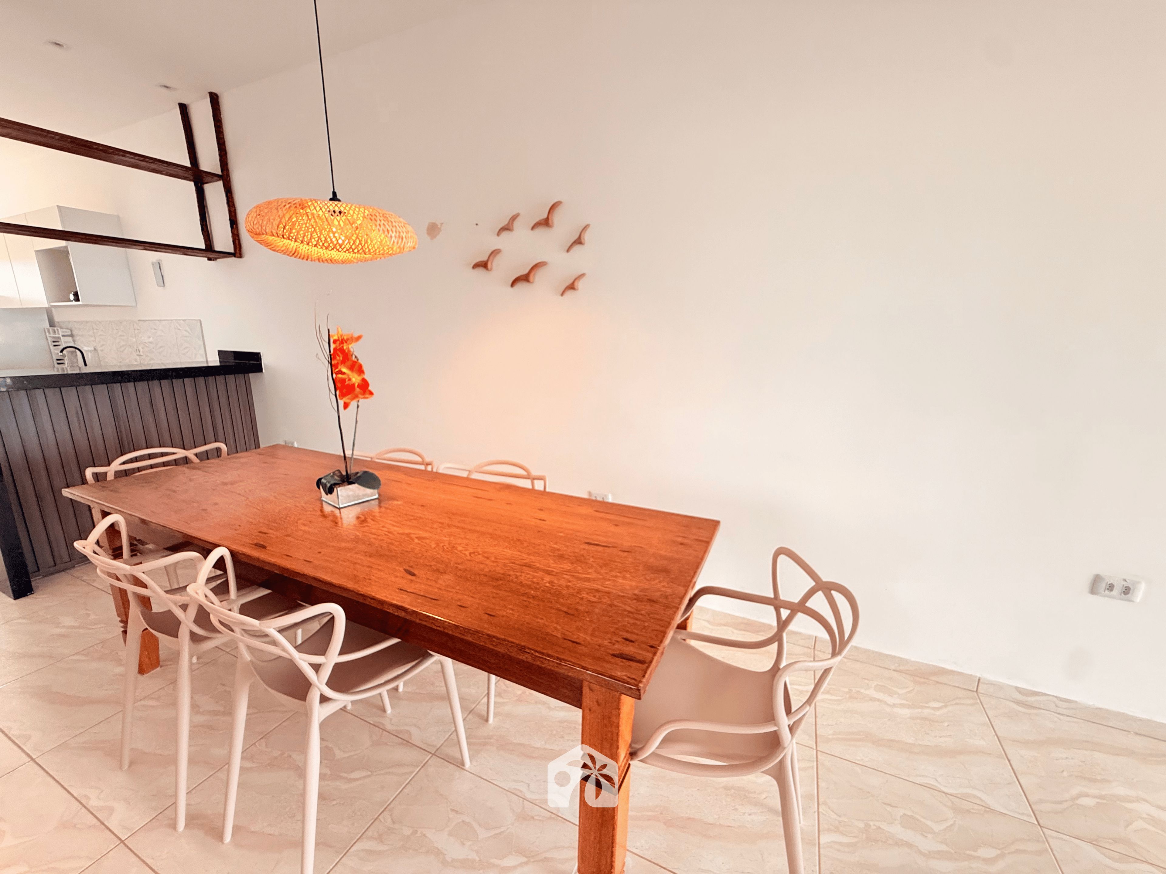 Un comedor con mesa de madera, sillas blancas, lámpara colgante y cocina al fondo.