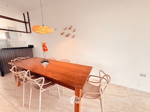 A dining area with a wooden table, white chairs, a hanging light fixture, and a kitchen in the background. - RoraiMar Residencial (Maragogi)