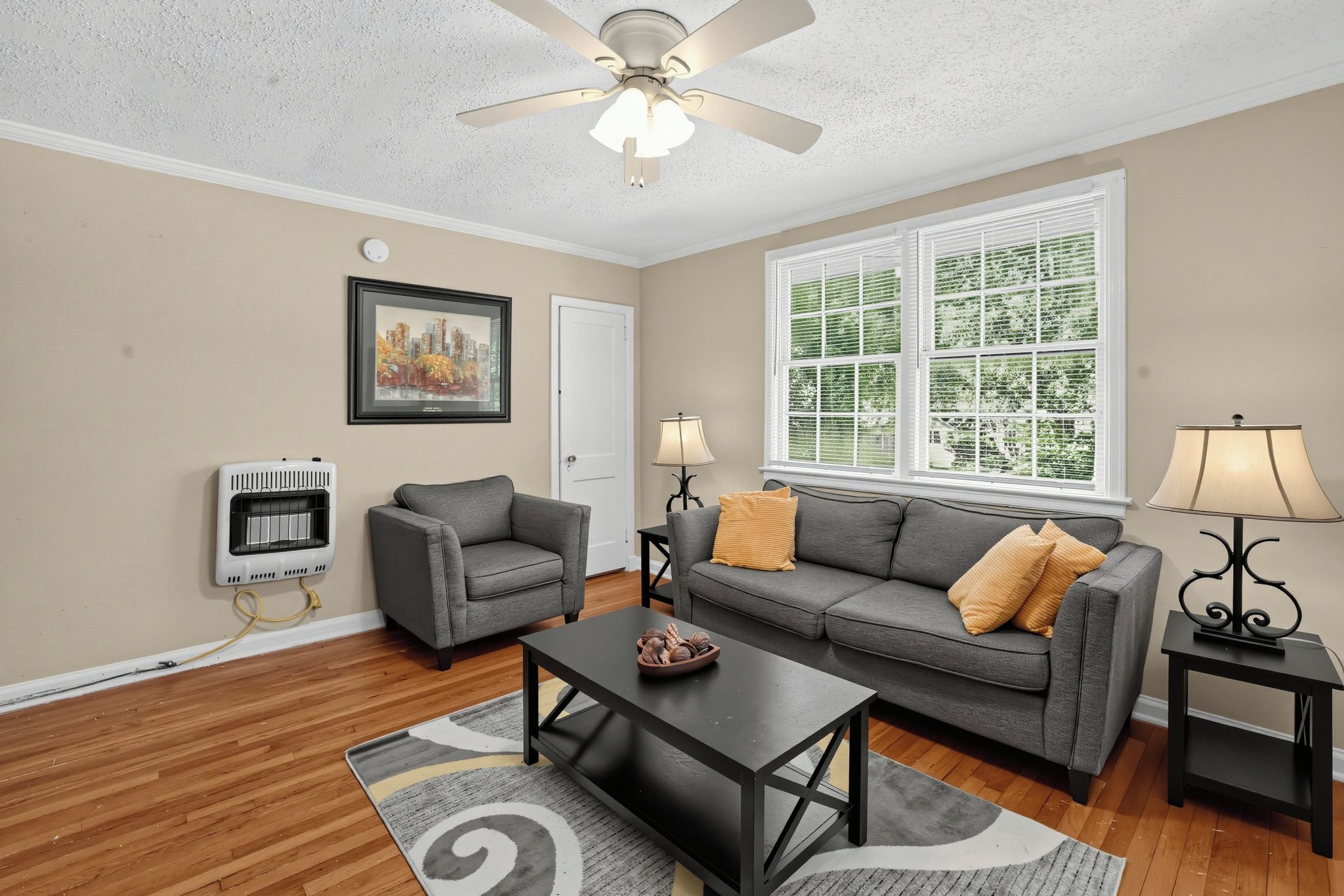 A living room with a gray sofa, a coffee table, a fireplace, a ceiling fan, and a window with blinds.