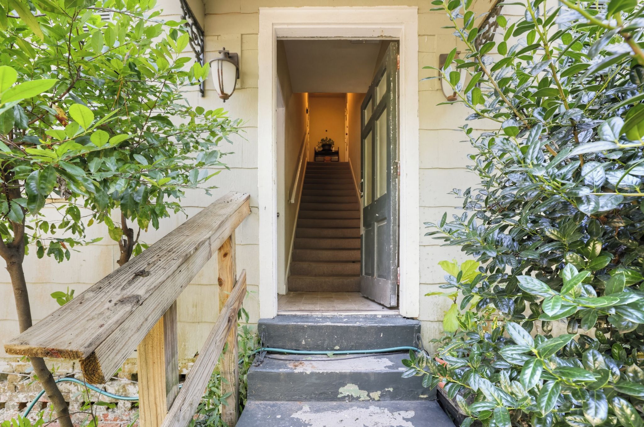 A wooden staircase leading to a door, surrounded by greenery and a wall-mounted light fixture.