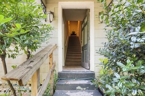 A wooden staircase leading to a door, surrounded by greenery and a wall-mounted light fixture.