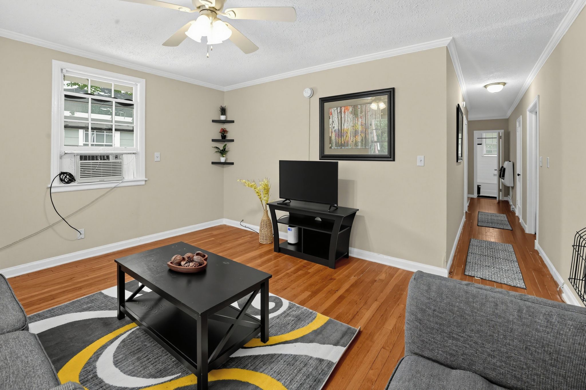 A living room with a flat-screen TV, a ceiling fan, a window with blinds, a fireplace, and a hallway leading to another room.