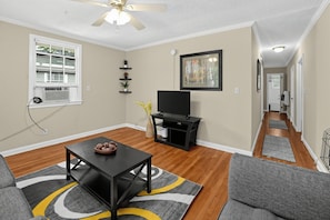 A living room with a flat-screen TV, a ceiling fan, a window with blinds, a fireplace, and a hallway leading to another room. - Old Cloverdale Cozy Nest (Montgomery)