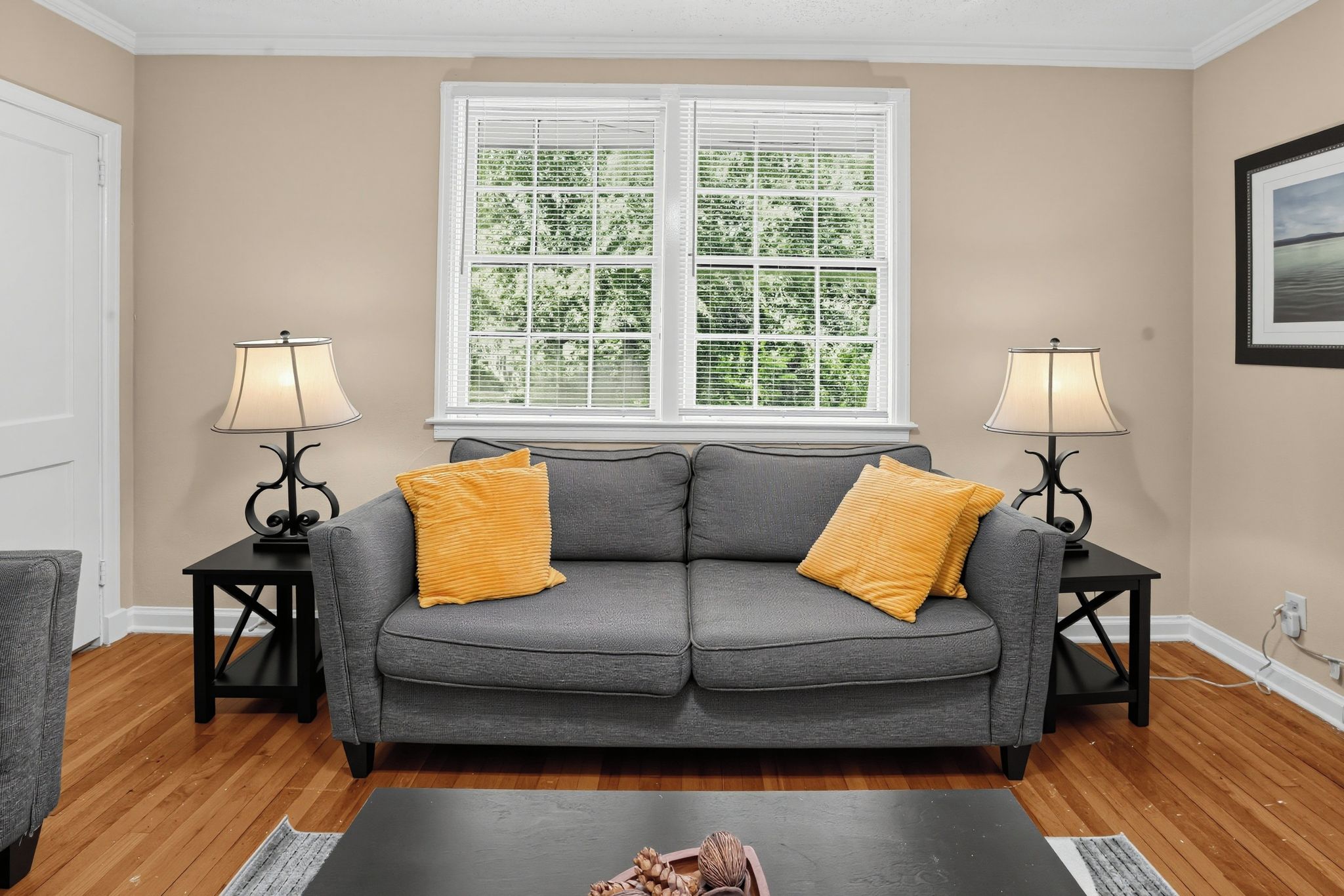 A living room with a grey sofa, two lamps, a window with blinds, and a framed picture on the wall.