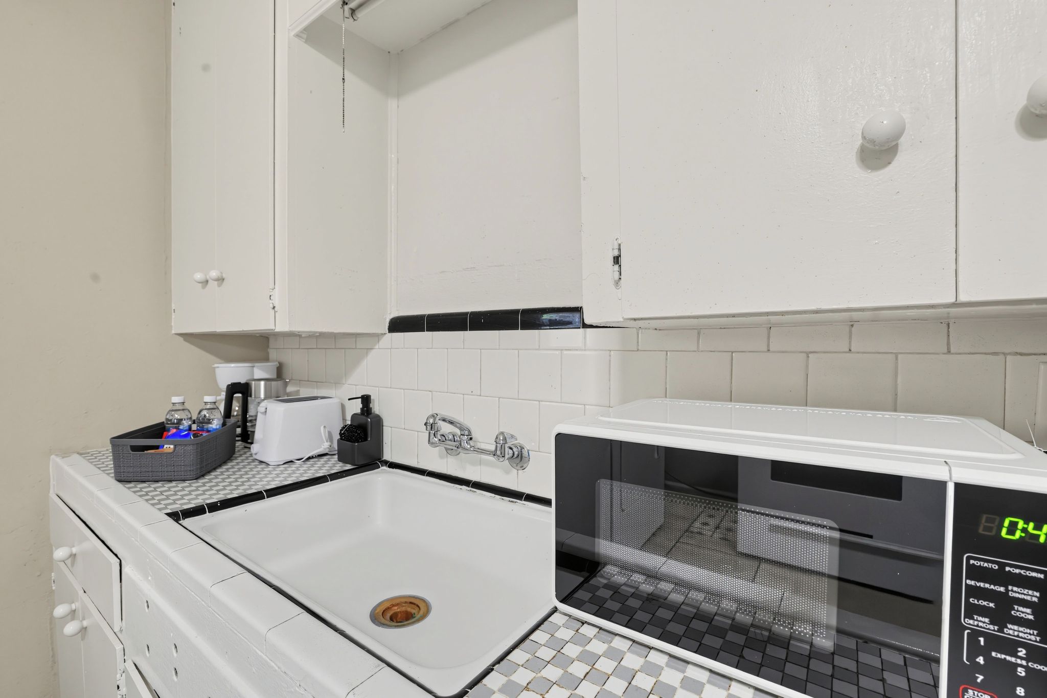 A kitchen with white cabinets, a microwave, a sink, and a small countertop with a kettle and a basket.