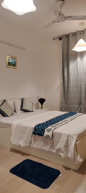 A bedroom with a bed, pillows, a bedside table, a lamp, a ceiling fan, and a window with curtains. - Spacious 2-BHK with AC close to Kempegowda international airport. (Bengaluru)