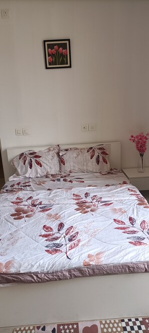 Ein Bett mit einer blumigen Tagesdecke, ein gerahmtes Bild an der Wand und eine Blumenvase auf einem Nachttisch.