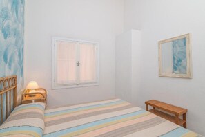 Ein Schlafzimmer mit einem Bett, einem Nachttisch mit Lampe, einem Fenster, einer Holzbank und einem gerahmten Bild an der Wand.