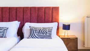 Ein Doppelbett mit weißer Bettwäsche und gemusterten Kissen, ein Nachttisch aus Holz mit einer Lampe sowie ein rotes, gepolstertes Kopfteil.