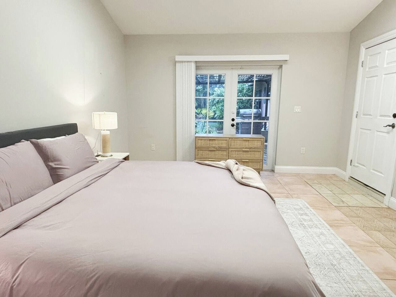 Quarto com cama, mesa de cabeceira, abajur, cômoda e porta que dá para uma varanda.