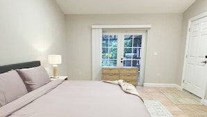 Ein Schlafzimmer mit Bett, Nachttisch, Lampe, Kommode und einer Tür zum Balkon.