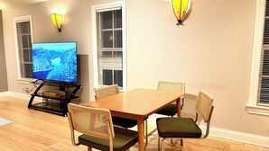 Een eetgedeelte met een houten tafel en vier stoelen, een televisie op een standaard en wandlampen.