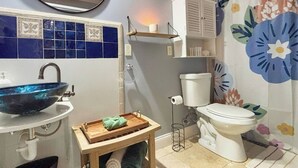 Ein Badezimmer mit blauen Fliesen, einer weißen Toilette, einem weißen Waschbecken mit blauer Schüssel, einem Holzregal mit einem weißen Behälter und einem blumigen Duschvorhang.