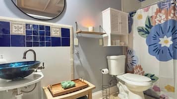 Um banheiro com parede em azulejos azuis, vaso sanitário branco, pia branca com cuba azul, prateleira de madeira com um recipiente branco e cortina de chuveiro florida.