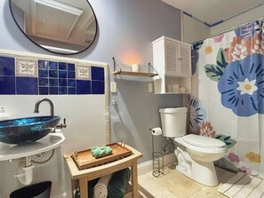 Um banheiro com parede em azulejos azuis, vaso sanitário branco, pia branca com cuba azul, prateleira de madeira com um recipiente branco e cortina de chuveiro florida.