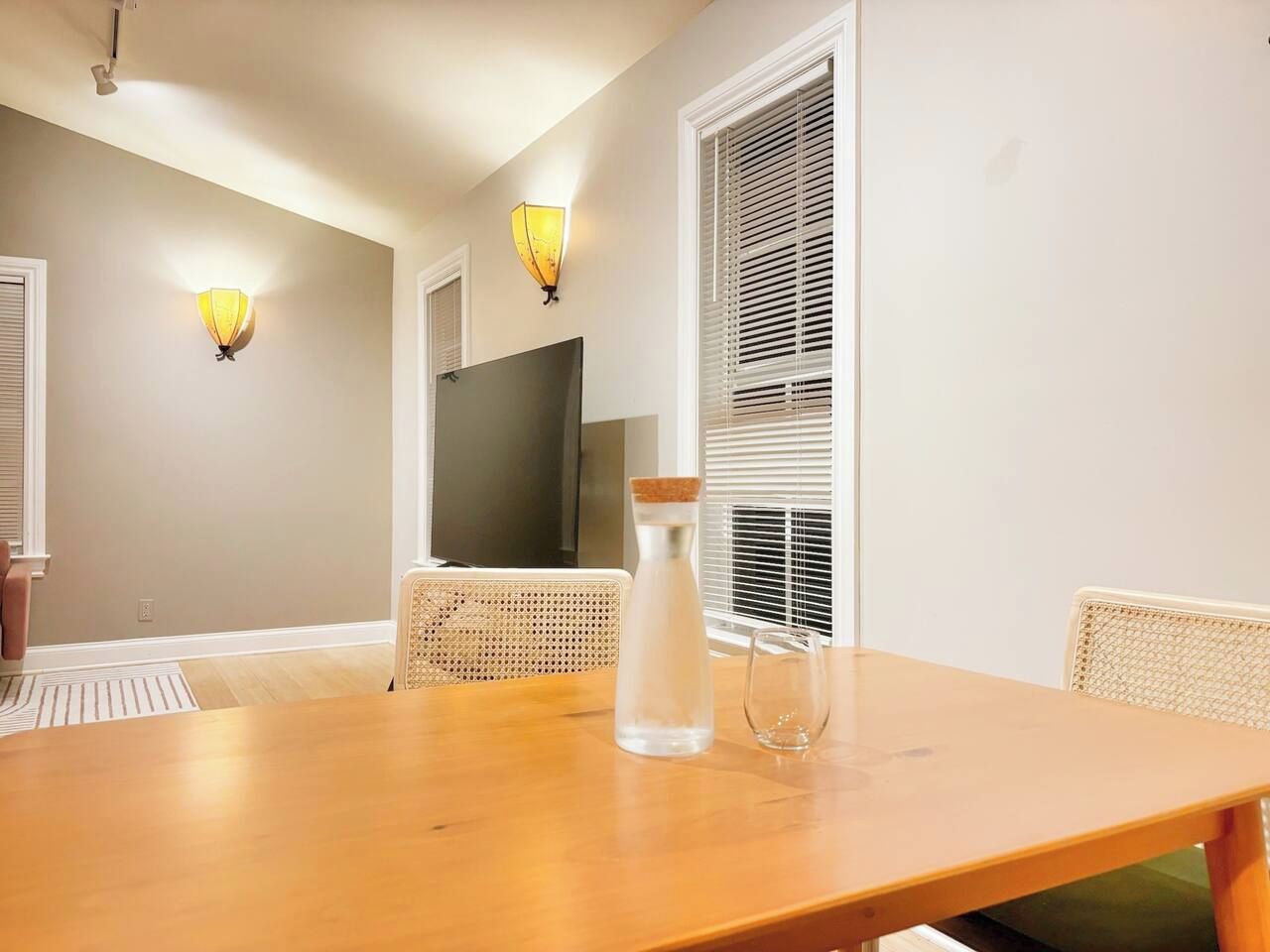 Uma área de refeições com uma mesa de madeira, duas cadeiras de vime, uma garrafa de água e duas luminárias fixadas na parede.
