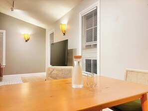 Uma área de refeições com uma mesa de madeira, duas cadeiras de vime, uma garrafa de água e duas luminárias fixadas na parede.