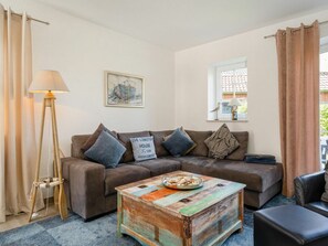 A cozy living room with a brown sofa, a colorful coffee table, a lamp, and a window with curtains. - Holiday Home in Greetsiel With Terrace (Krummhoern)