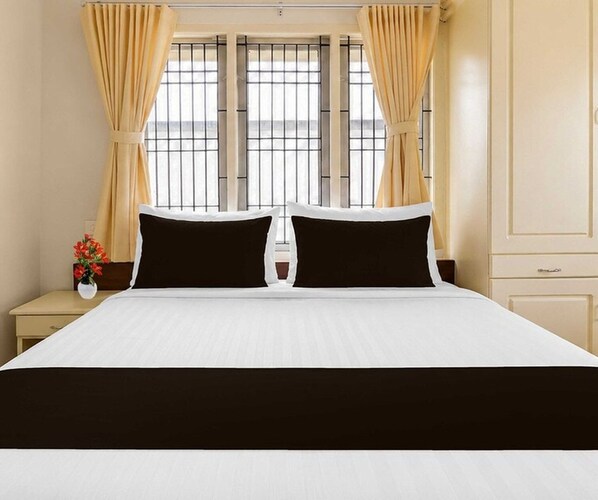 Una habitación de hotel con una cama grande, dos almohadas negras, una colcha blanca y cortinas color beige.