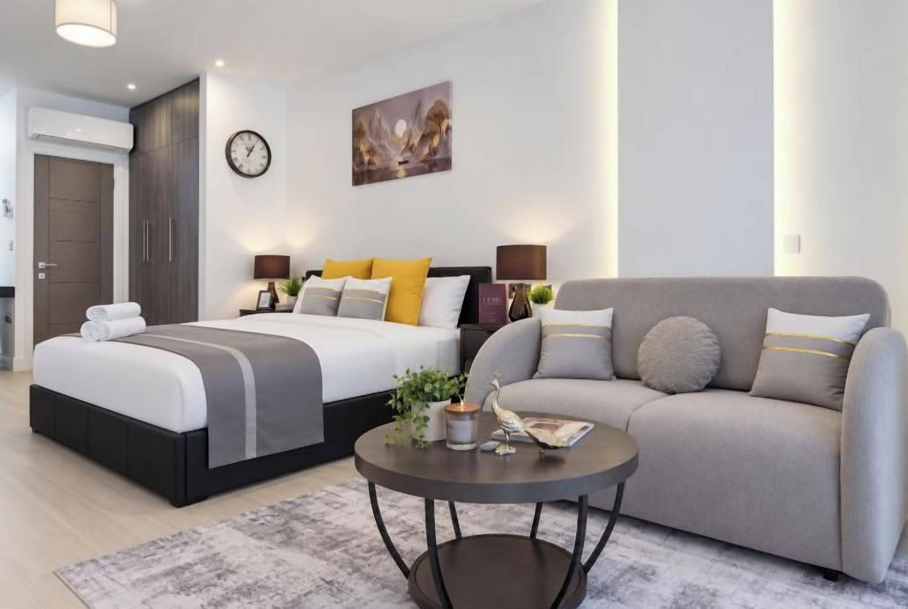 A modern hotel room with a double bed, a sofa, a coffee table, and a wall clock.