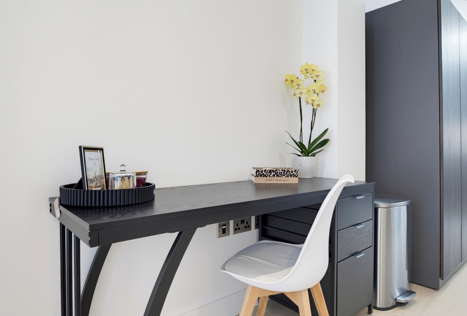 Un bureau moderne avec une chaise, une poubelle, un vase avec des fleurs jaunes et un tableau encadré posé sur le bureau.