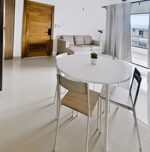 A modern living room with a round white table, two chairs, a small potted plant, a wooden door, a white sofa, and a view through a window with curtains. - The Reef · Pool & Home Cinema - Mins to Beach (Bayahíbe)