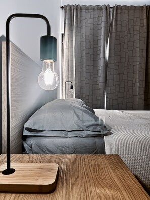 Een moderne slaapkamer met een houten nachtkastje, een lamp met een glazen kap, een grijs kussen, en een bed met grijze beddengoed.