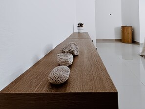 A long wooden table with four textured objects placed on it, a small potted plant, and a wooden trash bin in a minimalist room with white walls and a tiled floor. - The Reef · Pool & Home Cinema - Mins to Beach (Bayahíbe)