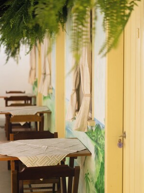 Una fila de mesas de madera con manteles estampados, una pared amarilla con un mural y una puerta con picaporte morado.