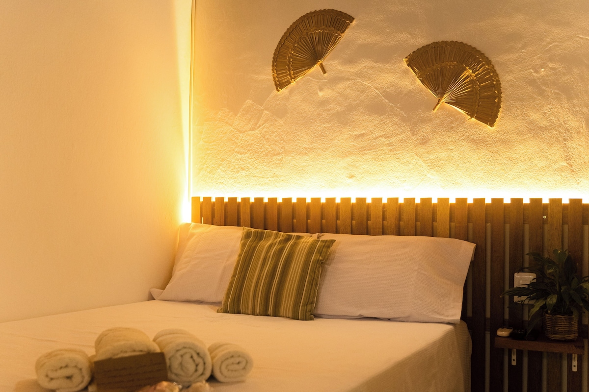 Ein ordentlich bezogenes Bett mit aufgerollten Handtüchern und einem Kopfkissen, ein Holz-Kopfteil und ein Wandventilator.