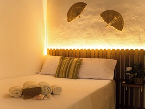 Ein ordentlich bezogenes Bett mit aufgerollten Handtüchern und einem Kopfkissen, ein Holz-Kopfteil und ein Wandventilator.