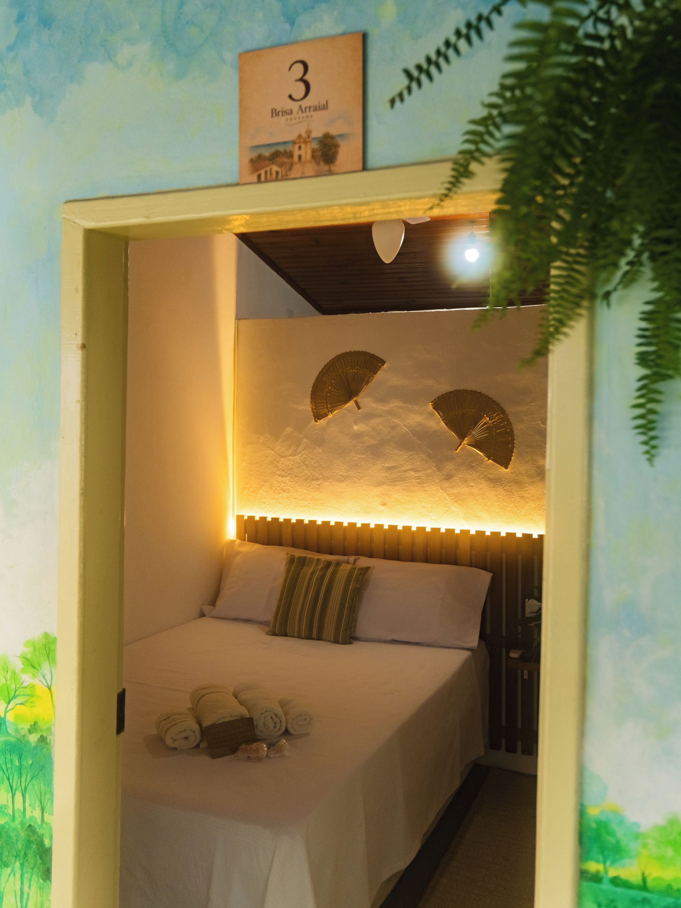 Quarto com cama, almofadas, ventoinha na parede e luminária no teto.
