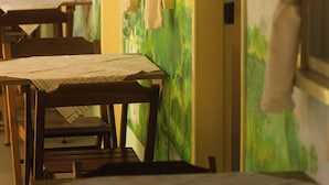 Quarto com mesas e cadeiras de madeira, um mural verde na parede e cortinas penduradas.