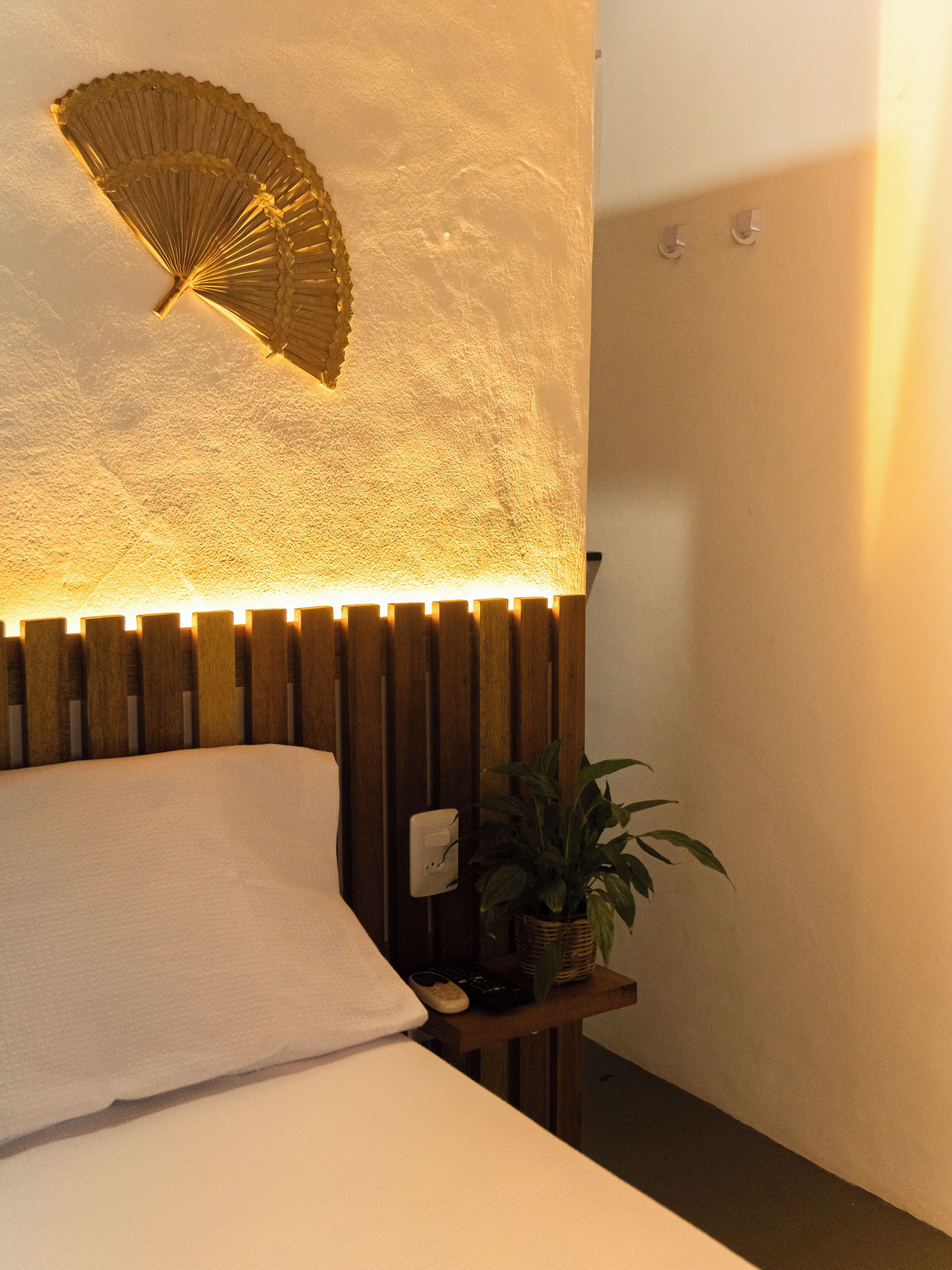 A bed with a white pillow, a wooden headboard, a wall-mounted fan, and a small table with a plant and a remote control.