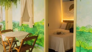 Quarto com cama, mesa, cadeiras e vista para uma paisagem pintada.