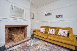 Un salon avec une cheminée, un canapé agrémenté de coussins à motifs floraux et des décorations murales portant les mots « PAIX » et « AMOUR ».